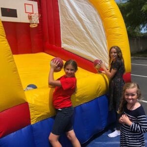 Three girls are enjoying a game with basketballs in front of an inflatable basketball setup. One girl, in a red shirt, holds a ball while the other two stand beside her, smiling brightly.