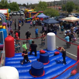 A lively crowd gathers outdoors for an event filled with inflatable bounce houses, games, and tents. Children eagerly navigate an inflatable obstacle course in the foreground.