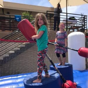 Two children stand on cushioned pedestals in a play area, gripping large red padded jousting sticks. They appear to be engaging in an inflatable jousting game with vibrant excitement. An adult is visible in the background.