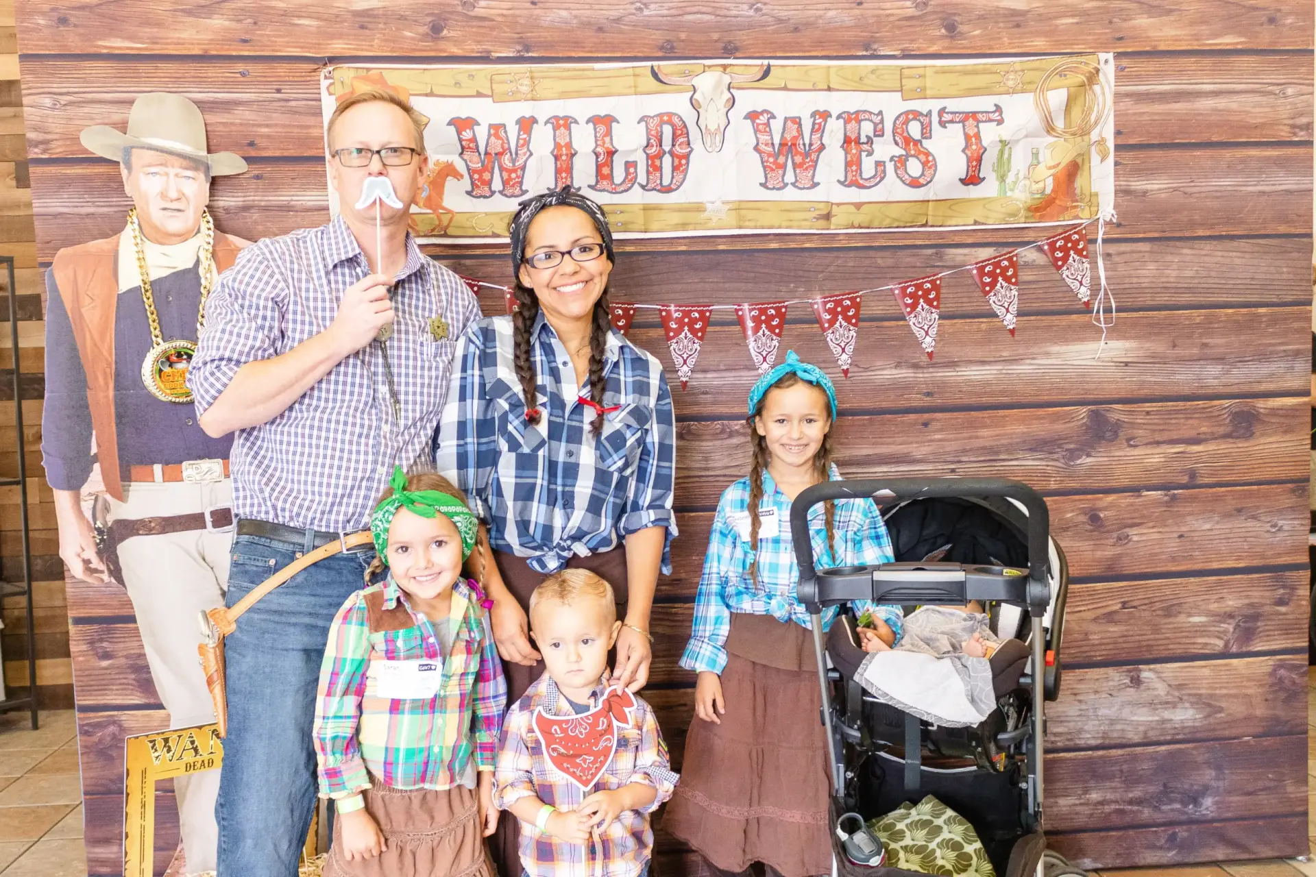 A family of five, dressed in Western-style clothing, stands in front of a "Wild West" sign with a wooden background. The father holds a mustache prop, the three children are smiling brightly, and a baby is tucked cozily in an antique stroller.