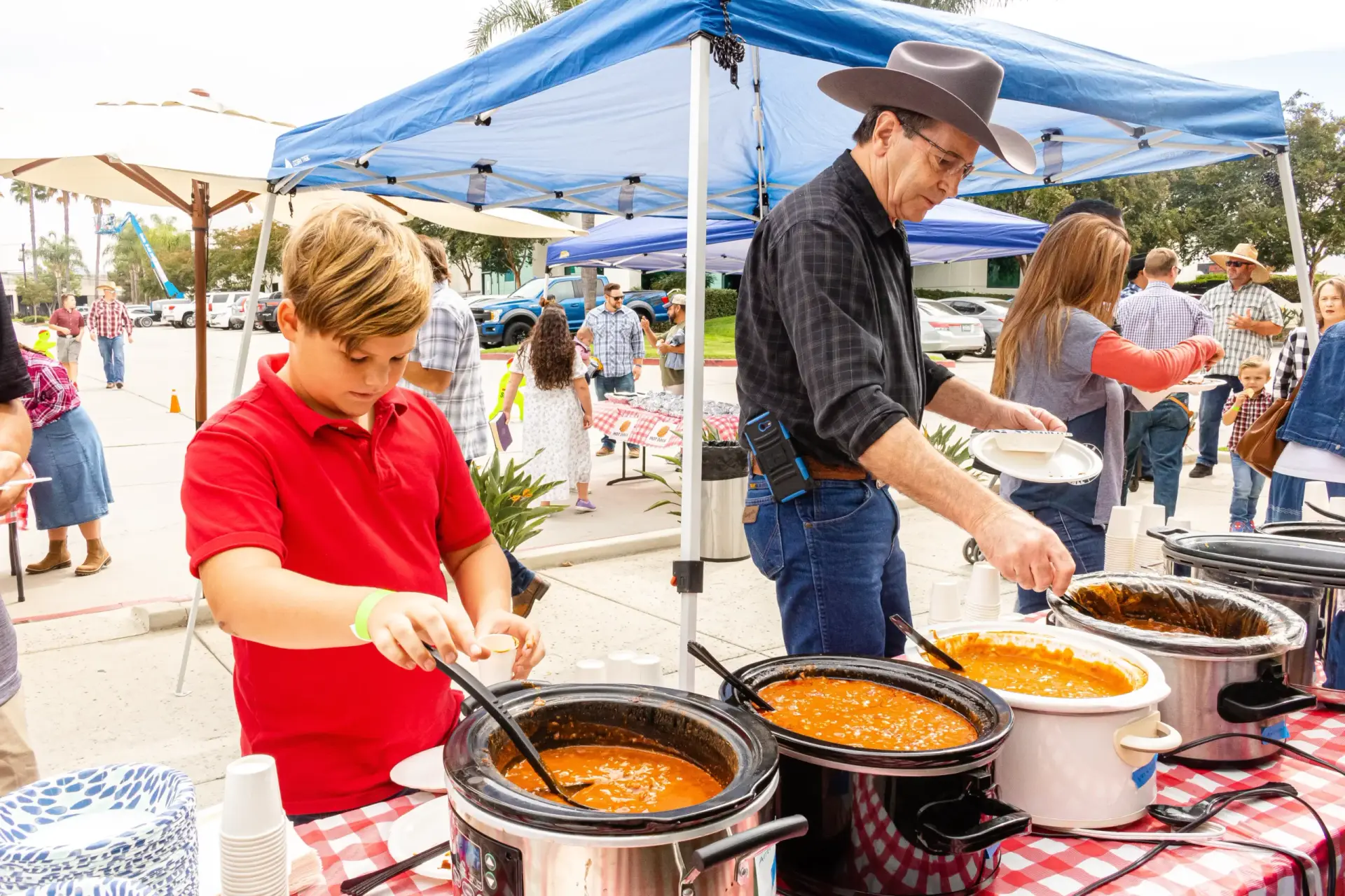 People serving themselves from crockpots under a blue canopy at an outdoor event. The table is adorned with a red and white checkered tablecloth, creating a cozy picnic atmosphere.