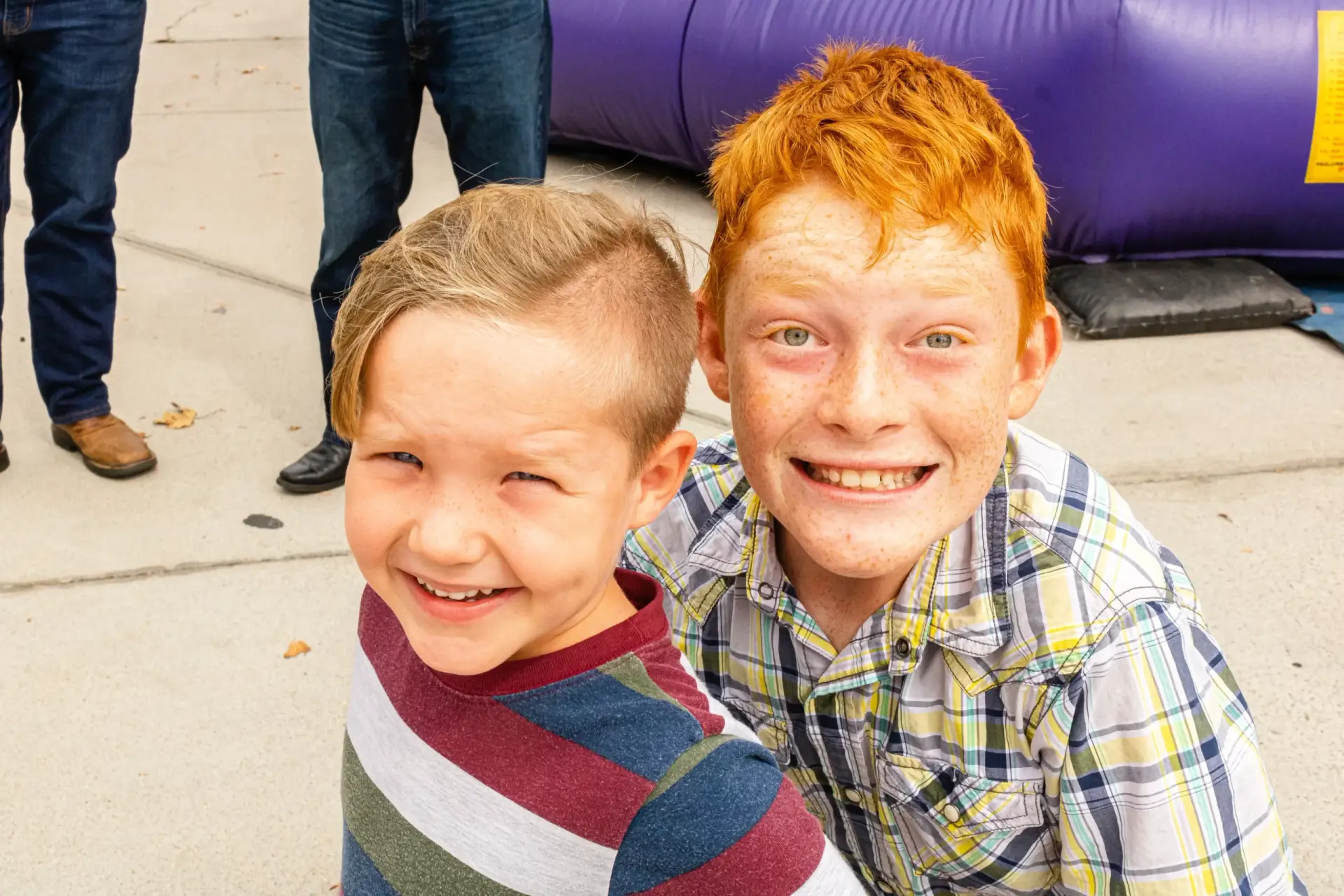 Two smiling children, one with red hair in a plaid shirt and the other with blonde hair in a striped shirt, standing outdoors on the sidewalk.