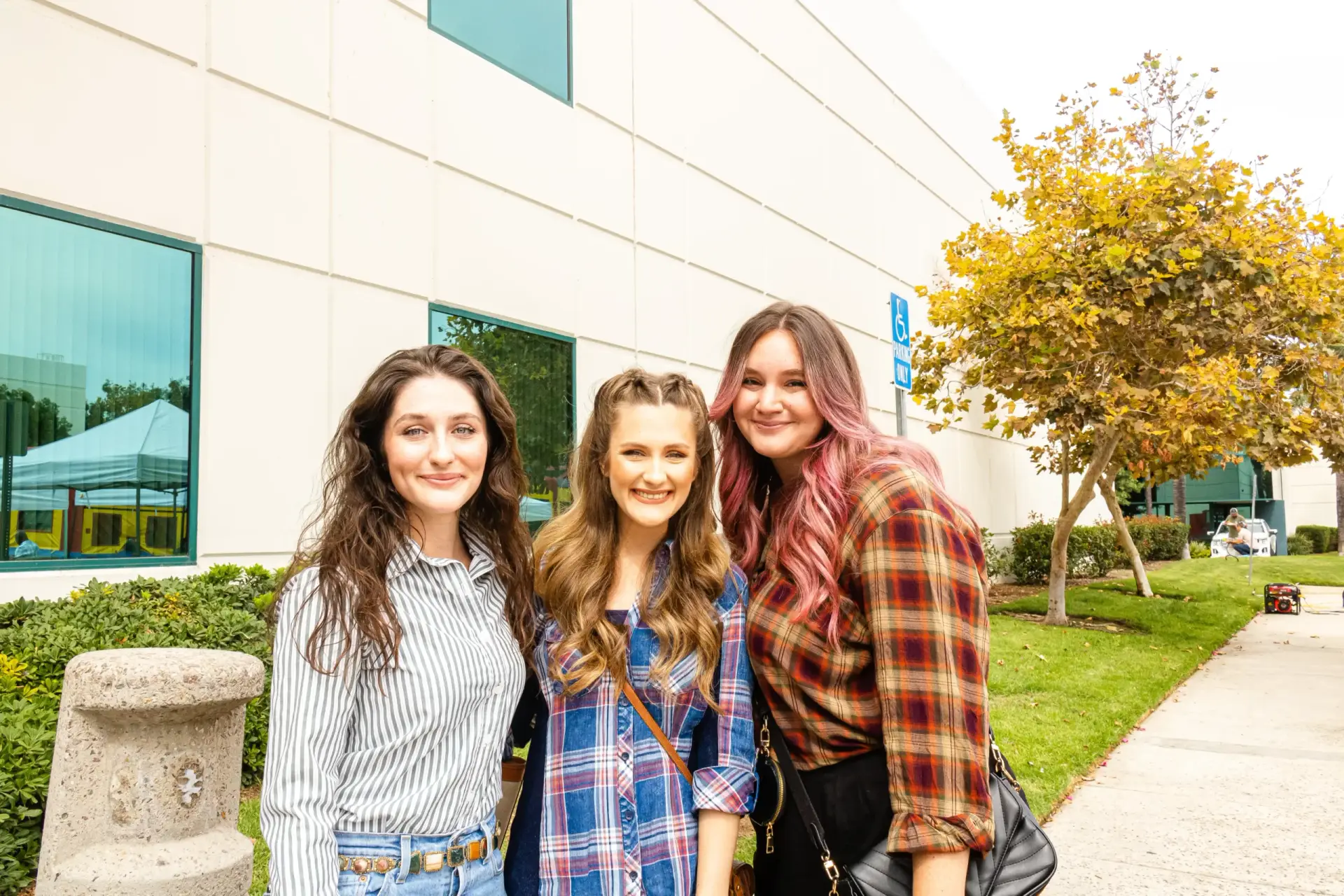 Three young women stand close together, smiling in front of a modern beige building with large windows. One has dark hair, the second light brown hair, and the third sports pink highlights.