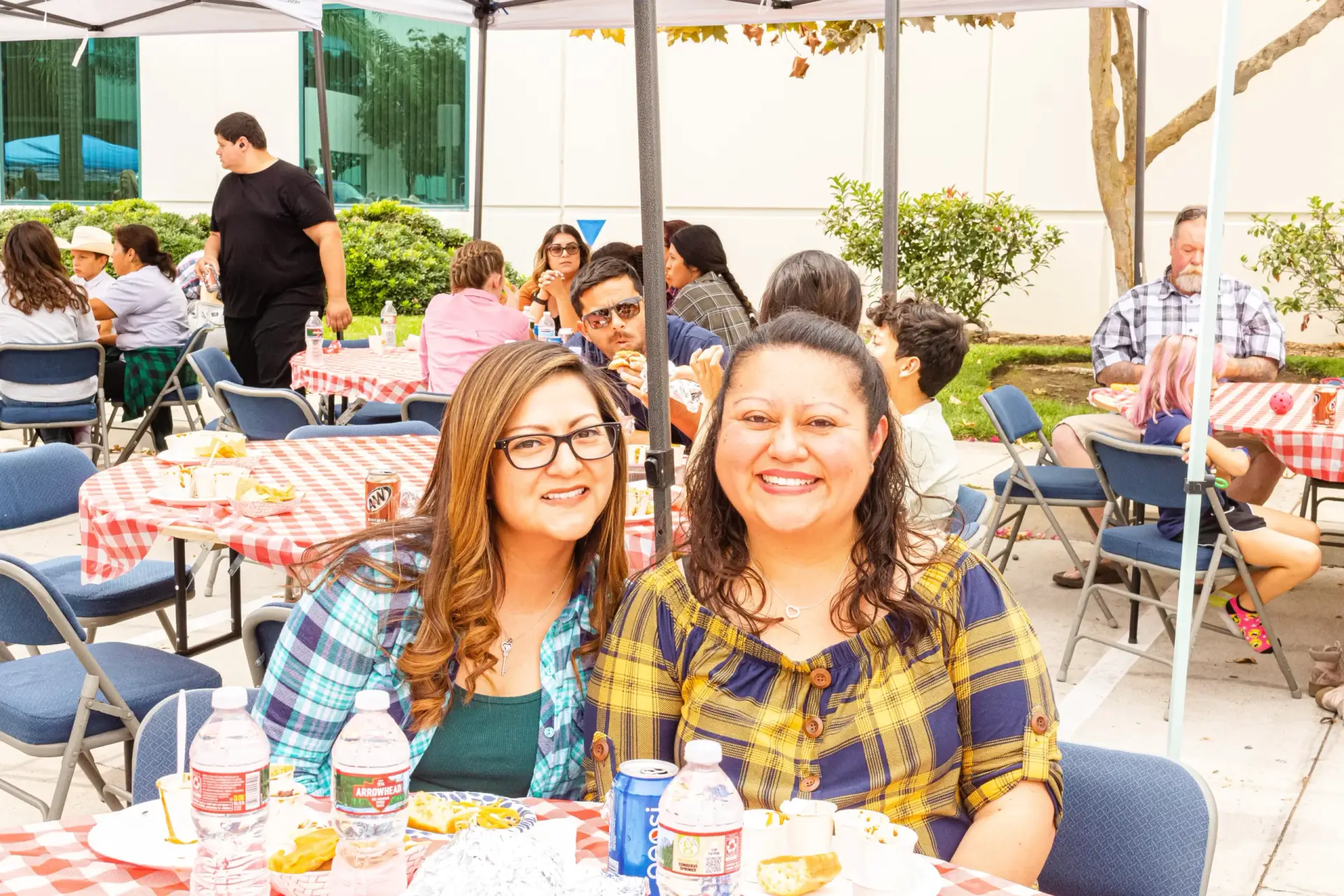 Two women smile for the camera while sitting at a picnic table with a red checkered tablecloth, surrounded by other people enjoying the outdoor setting at similar tables.