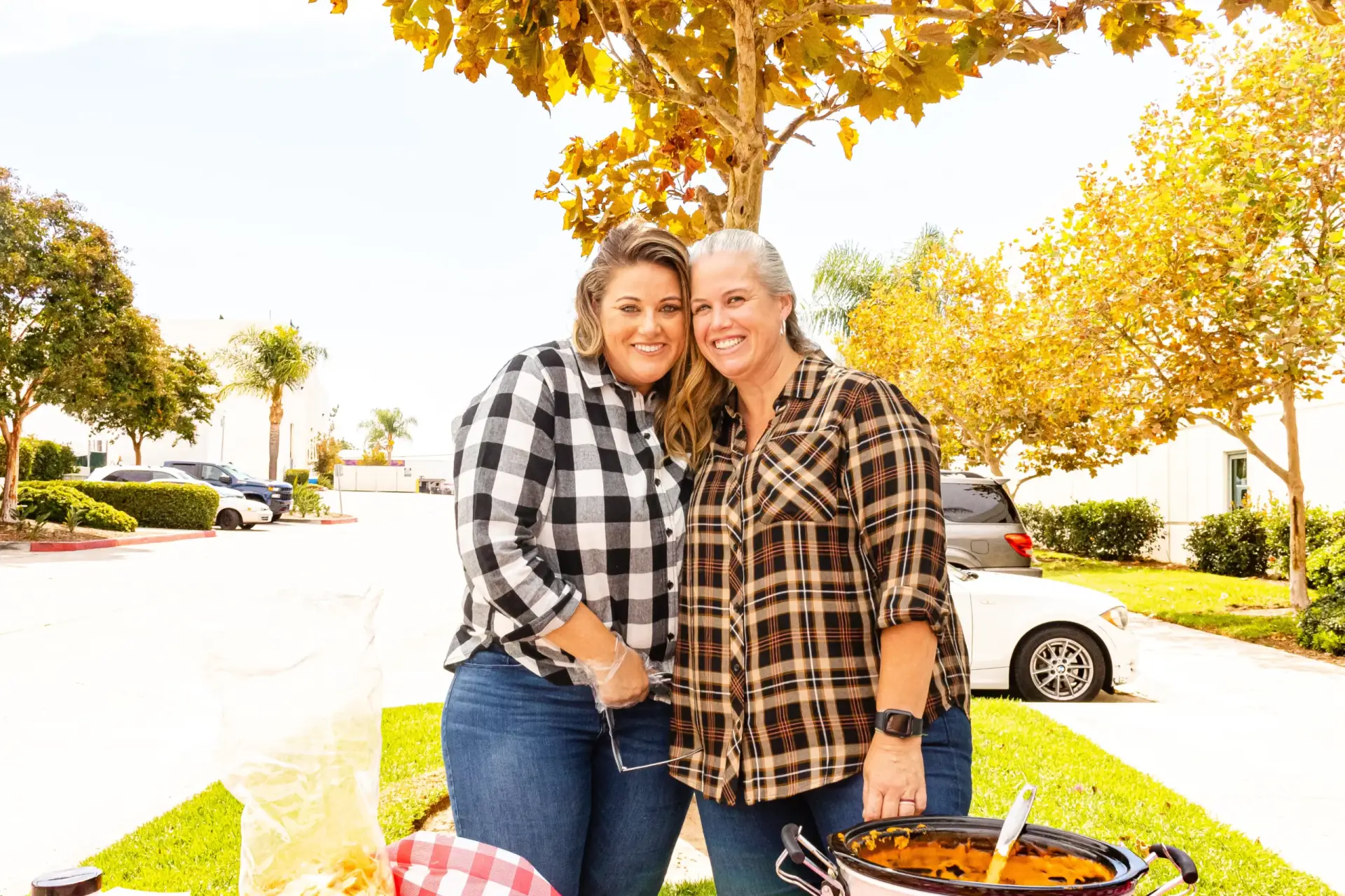 Two smiling people stand close together outdoors, with a table of delicious food in front of them. They are dressed in casual clothes, and trees with vibrant autumn foliage are in the background, adding a touch of seasonal beauty to the scene.