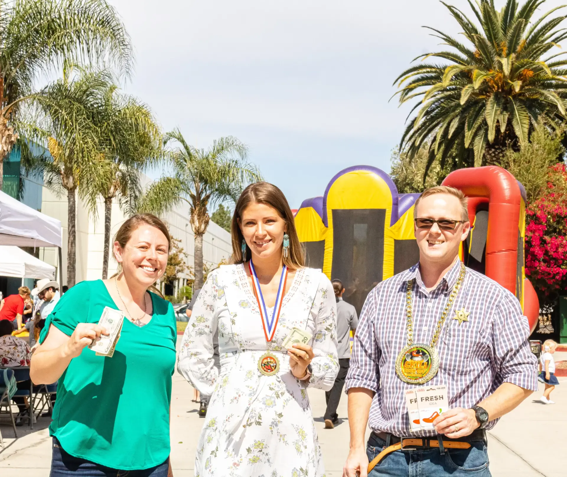 Three people stand outdoors holding awards. The woman in the center wears a medal. Palm trees sway gently in the background while an inflatable bounce house adds a fun touch to the scene.