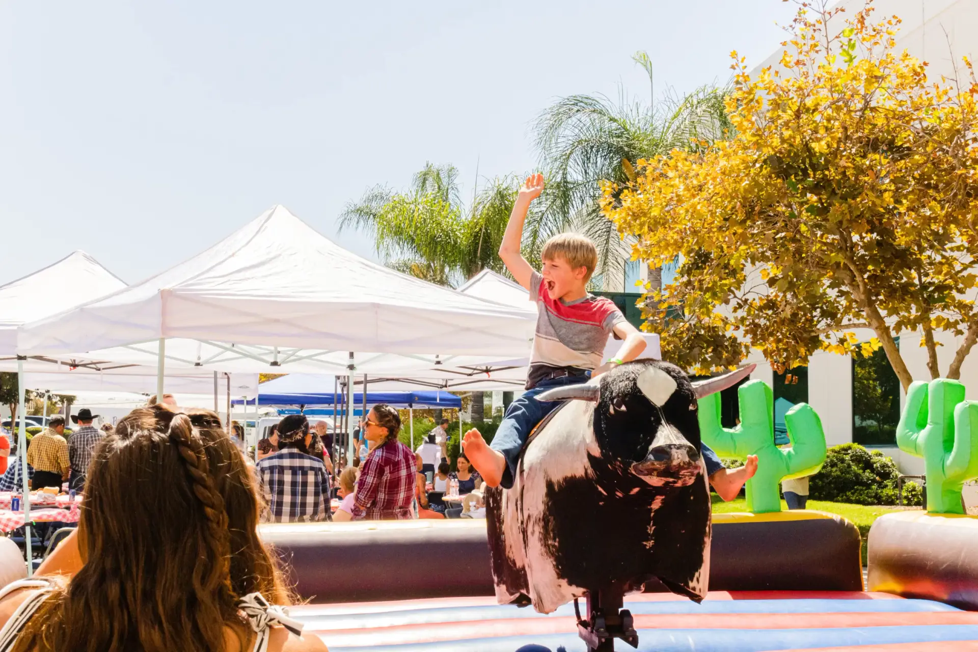 A child, holding up one arm, rides a mechanical bull in an outdoor setting with tents, palm trees, and festive music playing in the background.