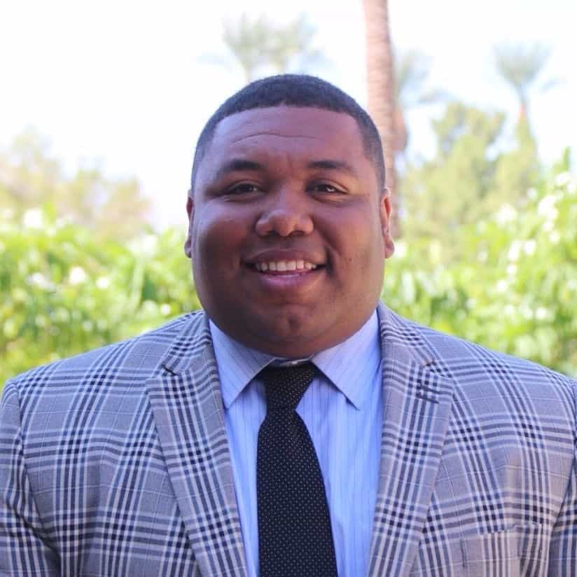 A man in a checked suit jacket, light blue shirt, and black tie smiles outdoors with lush greenery in the background.