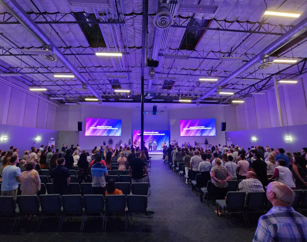 Audience standing and watching a live band perform on stage at a spacious indoor event hall with vibrant lighting and large video screens, during a Worship Service at a Church in El Cajon.